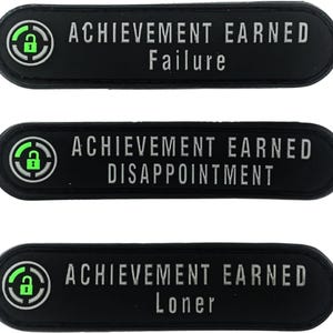 Może przedstawiać: Trzy czarne, owalne naszywki z białym tekstem. Każda naszywka zawiera napis "ACHIEVEMENT EARNED" nad innym słowem: "Failure", "Disappointment" i "Loner". Każda naszywka ma zielono-białą grafikę kłódki.