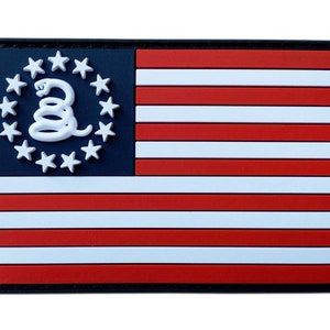 Könnte beinhalten: Ein schwarz-weißes Patch mit einem rot-weiß-blauen amerikanischen Flaggen-Design. Die Flagge hat im oberen linken Eck einen Kreis aus Sternen mit einer darin eingerollten Schlange. Die Schlange ist weiß und hat einen eingerollten Schwanz. Der Text "Don't Tread On Me" ist auf dem Bild nicht sichtbar.