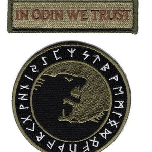 Puede incluir: Dos parches bordados. El parche superior es un parche rectangular verde oliva con el texto "IN ODIN WE TRUST" en letras marrones. El parche inferior es un parche redondo negro con un círculo blanco alrededor del borde. El círculo blanco tiene runas blancas alrededor del borde. Dentro del círculo hay una cabeza de lobo negra y verde oliva.
