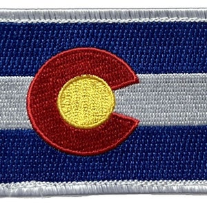 May include: An embroidered patch featuring the state flag of Colorado. The flag is blue, white and red with a large 'C' in the centre, with a gold circle inside.