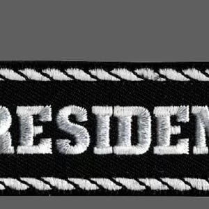 Könnte beinhalten: Schwarz-weißer gestickter Aufnäher mit weißem Kordelrand. Der Aufnäher trägt die Aufschrift "PRESIDENT" in weißen Buchstaben.