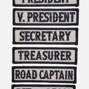 Może przedstawiać: Sześć białych naszywek haftowanych czarnym szwem. Naszywki przedstawiają napisy: "Prezes", "Wiceprezes", "Sekretarz", "Skarbnik", "Kapitan Drogi" i "Sztandarowy".