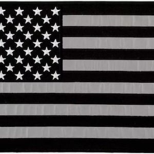 Puede incluir: Un parche de bandera estadounidense en blanco y negro con un acabado reflectante. Las estrellas son blancas y las rayas son grises.