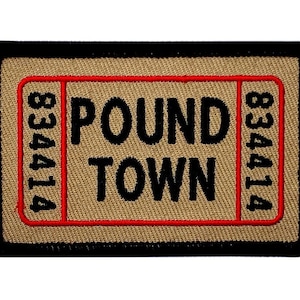 Può includere: Una toppa ricamata di colore beige e nero con cuciture rosse. La toppa ha la forma di un biglietto e dice "POUND TOWN" con il numero "834414" su entrambi i lati.