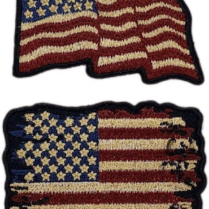 May include: Two embroidered American flag patches. The top patch is a waving flag with red and white stripes and gold stars on a blue field. The bottom patch is a distressed flag with red and white stripes and gold stars on a blue field. The edges of the bottom patch are frayed.