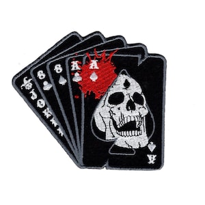 May include: Black and white embroidered patch of a skull with a red blood splatter behind it. The skull is wearing a black ace of spades playing card. The patch is surrounded by other playing cards, including the 8 of spades, the jack of spades, and the joker.
