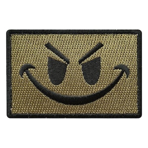 Könnte beinhalten: Gestickter Aufnäher mit goldenem Hintergrund und schwarzen Details, der ein schelmisches Smiley-Gesicht darstellt. Der rechteckige Aufnäher hat einen schwarzen Rand. Die Augen sind nach oben gerichtet und der Mund formt ein finsteres Grinsen.