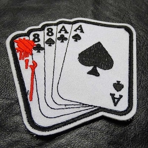 May include: A white embroidered patch with five playing cards, four of which are aces of spades and one is an eight of spades. The cards are arranged in a fan shape with the eight of spades on the bottom and the aces of spades on top. The patch has a black border.