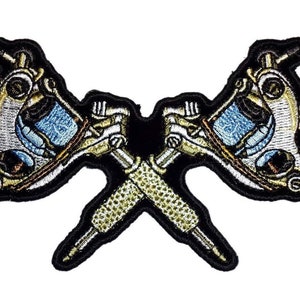 Puede incluir: Un parche bordado en blanco y negro de dos máquinas de tatuaje cruzadas. Las máquinas están detalladas con detalles dorados y azules.