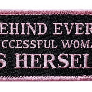以下が含まれることがあります： ピンクのステッチで刺繍された黒いパッチ。テキストは「Behind every successful woman is herself.」です。
