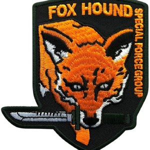 Puede incluir: Un parche bordado negro y verde con una cabeza de zorro y un cuchillo. El texto "FOX HOUND SPECIAL FORCES GROUP" está en el parche.