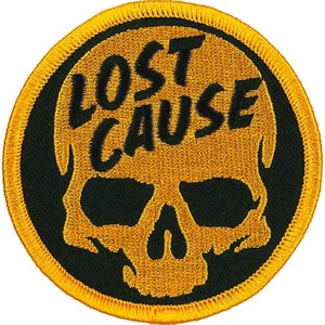 Könnte beinhalten: Runder, gestickter Aufnäher mit einem goldenen Totenkopf-Design und dem Schriftzug "LOST CAUSE" in Schwarz. Der Totenkopf befindet sich auf einem schwarzen Hintergrund, mit einem goldenen Rand. Geeignet für Kleidung oder Accessoires.