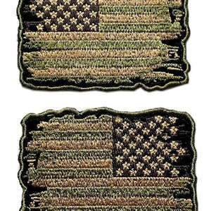 Może przedstawiać: Dwa haftowane naszywki z amerykańską flagą w kamuflażu. Naszywki są strzępione na krawędziach, co nadaje im zużyty wygląd.