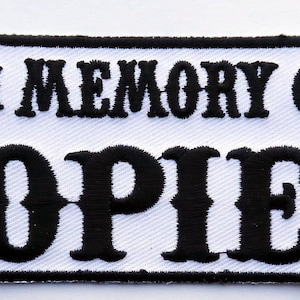 Op de afbeelding: Witte geborduurde patch met zwarte tekst "In Memory of 'Opie'".