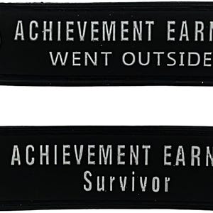 Könnte beinhalten: Zwei schwarze, ovale Aufnäher mit weißem Text. Der obere Aufnäher lautet "ACHIEVEMENT EARNED" und "WENT OUTSIDE". Der untere Aufnäher lautet "ACHIEVEMENT EARNED" und "SURVIVOR". Jeder Aufnäher hat eine grün-weiße Grafik eines Schlosses.