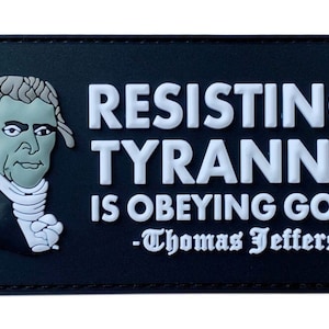 Può includere: Toppa bianca e nera con un'immagine di cartone animato di Thomas Jefferson e il testo "Resistere alla tirannia è obbedire a Dio. -Thomas Jefferson"