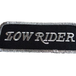 Op de afbeelding: Een zwarte geborduurde patch met zilverdraad, met de tekst "LOW RIDER".
