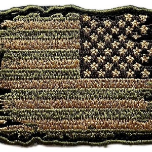 May include: An embroidered patch of the American flag in green and brown colours, with a distressed look. The flag is frayed around the edges and has a worn appearance.