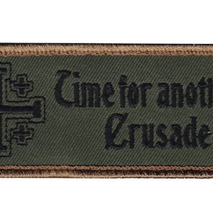Peut inclure: Un écusson brodé vert avec une bordure marron. L'écusson représente une croix noire avec un centre blanc et le texte "Time for another Crusade".