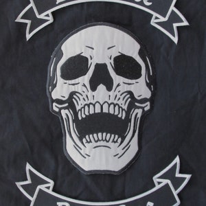 Pode incluir: Patch bordado em preto e branco com um design de caveira e ossos cruzados. O patch tem duas faixas com o texto "Live Free" e "Ride Free".