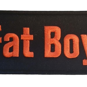 Może przedstawiać: Czarny prostokątny znaczek z napisem "Fat Boy" w haftowanych pomarańczowych literach.