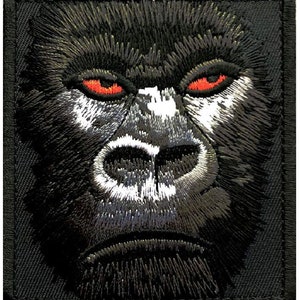 May include: Close-up of a black and white embroidered gorilla face patch with red eyes. The patch is on a black background.