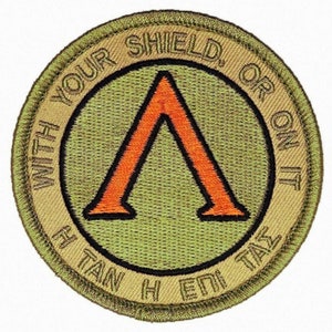 May include: A green and tan embroidered patch with a black outline. The patch features a red and orange stylized letter A with the text "With your shield, or on it" around the outside of the A. The text "H E DI TAN H" is around the outside of the patch.