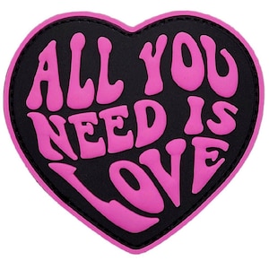 Pode incluir: Um remendo em forma de coração rosa com o texto "ALL YOU NEED IS LOVE" em uma fonte retrô.