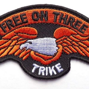 Puede incluir: Parche bordado naranja y negro con una pancarta que dice "Free On Three" y un águila blanca con alas. La parte inferior del parche dice "Trike".