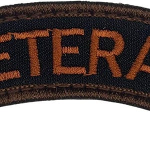 May include: A curved patch with the word "VETERAN" embroidered in orange thread on a navy blue background. The patch has a brown border and is likely designed to be sewn onto clothing or gear.