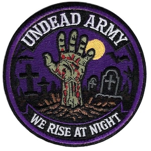 May include: A circular embroidered patch featuring a green zombie hand emerging from the ground in a graveyard at night. The design includes a yellow moon, black silhouettes of tombstones, and the text "UNDEAD ARMY" and "WE RISE AT NIGHT" in white.