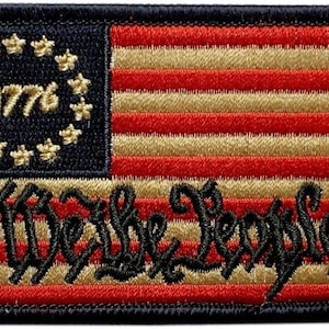 以下が含まれることがあります： 13本のストライプと星の円を持つ、スタイリッシュなアメリカ国旗が刺繍された黒と金のワッペン。旗の下部に「We the People」という文字が黒で刺繍されています。