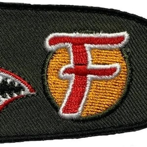 May include: An embroidered patch shaped like a bomb in olive green, featuring a shark mouth graphic with white teeth and red accents. A yellow circle contains a large red letter F. The design includes a black border and a finned tail section.