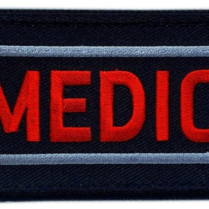 Puede incluir: Un parche negro con dos rayas blancas y la palabra "MEDIC" en letras bordadas rojas.
