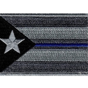 Könnte beinhalten: Ein gestickter Patch mit dem Design der puerto-ricanischen Flagge in Schwarz und Weiß. Die Flagge hat einen blauen Streifen in der Mitte und einen weißen Stern im schwarzen Dreieck. Der Patch hat einen grauen Rand.