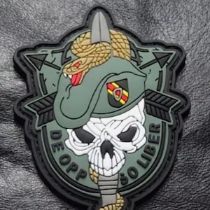 Puede incluir: Un parche bordado verde, negro y blanco con una calavera, una serpiente y una boina. El texto "De Oppresso Liber" está escrito alrededor de la calavera.