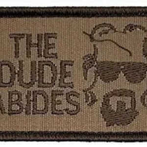 Pode incluir: Um remendo bordado marrom com fundo bege. O remendo apresenta uma imagem de desenho animado de um homem com óculos de sol e barba. O texto "THE DUDE ABIDES" é bordado em marrom no remendo.