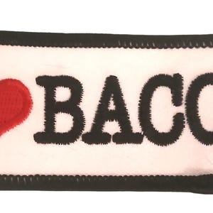 Puede incluir: Un parche rectangular con las palabras "I ❤️ BACON" bordadas en negro sobre un fondo blanco. Un corazón rojo reemplaza la palabra "love". El parche tiene un borde negro.