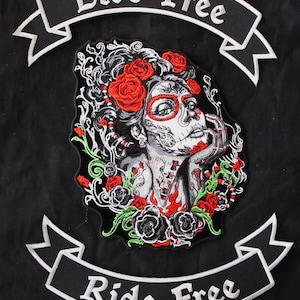 Może przedstawiać: Haftowana naszywka z kobietą w stylu sugar skull, czerwonymi różami i napisem "Live Free Ride Free" na czarnym tle. Twarz kobiety zdobią skomplikowane wzory, a róże są w żywym kolorze.
