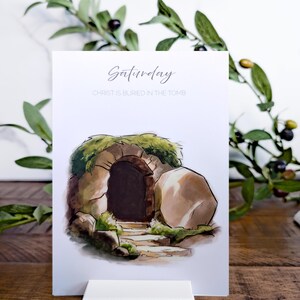 May include: A white card with a watercolor illustration of a stone tomb with a large stone rolled away. The text on the card reads "Saturday" and "Christ is buried in the tomb". The card is standing on a white card stand.