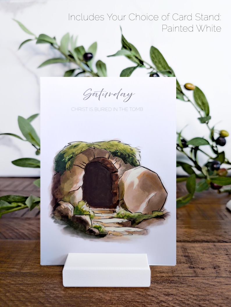 May include: A white card with a watercolor illustration of a stone tomb with a large stone rolled away. The text on the card reads "Saturday" and "Christ is buried in the tomb". The card is standing on a white card stand.