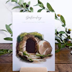 May include: A white card with a watercolor illustration of a stone tomb with a large stone rolled away. The text "Saturday" and "CHRIST IS BURIED IN THE TOMB" is printed on the card. The card is standing on a small wooden stand.