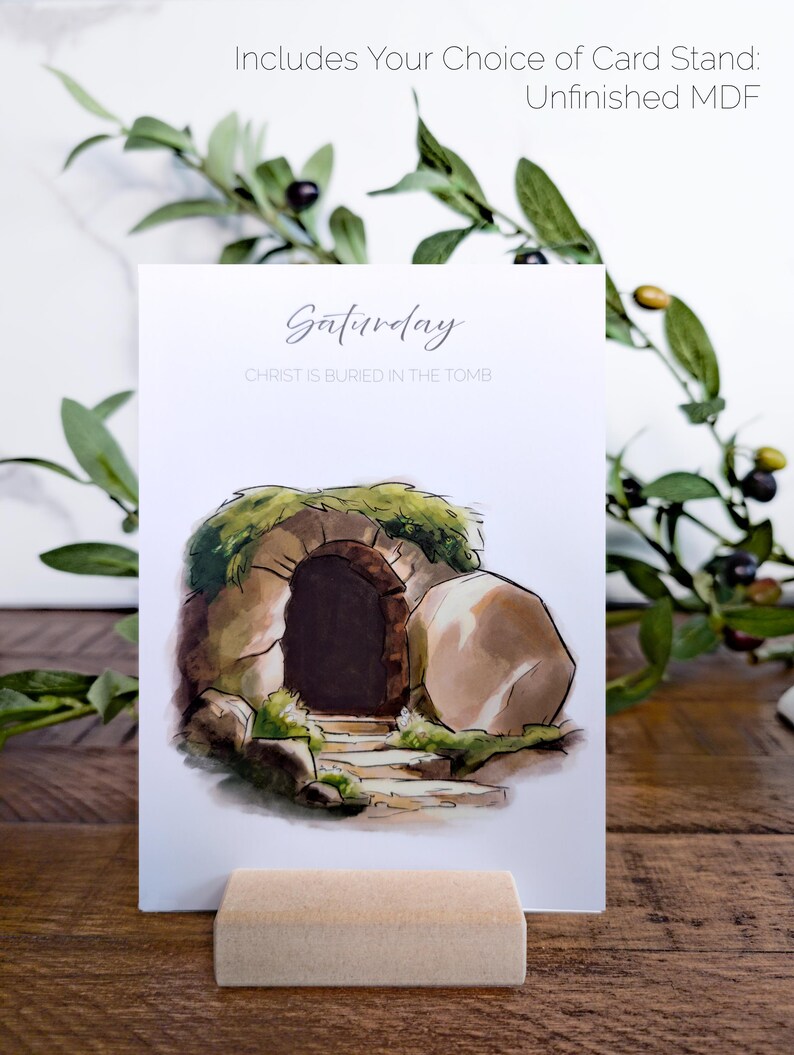 May include: A white card with a watercolor illustration of a stone tomb with a large stone rolled away. The text "Saturday" and "CHRIST IS BURIED IN THE TOMB" is printed on the card. The card is standing on a small wooden stand.
