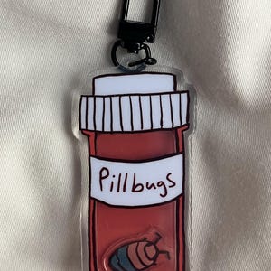 May include: A clear acrylic keyring featuring a red pill bottle design with the text "Pillbugs" on a white label. Inside the bottle is a cartoon pillbug. It has a black clasp.