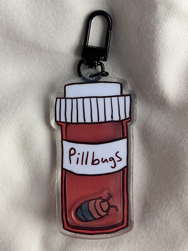 May include: A clear acrylic keyring featuring a red pill bottle design with the text "Pillbugs" on a white label. Inside the bottle is a cartoon pillbug. It has a black clasp.