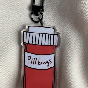 May include: A clear acrylic keyring with a red pill bottle design, a white label, and the text "Pillbugs". The keyring has a black metal clasp for attaching to keys or bags.