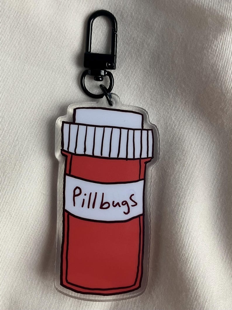 May include: A clear acrylic keyring with a red pill bottle design, a white label, and the text "Pillbugs". The keyring has a black metal clasp for attaching to keys or bags.