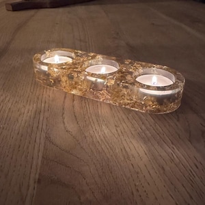 May include: A clear glass candle holder with three compartments for tea lights. The holder is decorated with gold leaf and has a rustic wooden background.