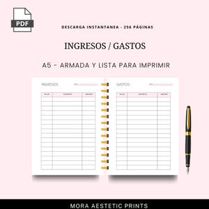 Puede incluir: Un planificador imprimible A5 rosa y blanco con encuadernaci&oacute;n en espiral dorada. El planificador est&aacute; abierto en dos p&aacute;ginas con secciones para ingresos y gastos. Un bol&iacute;grafo negro y dorado descansa en el lado derecho. El texto incluye "INGRESOS / GASTOS".