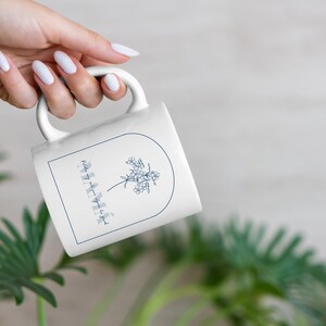 Pode incluir: Caneca de cer&acirc;mica branca com um design floral azul e o texto "MAMA" com as palavras "You make my world a better place" abaixo.